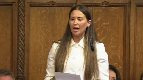 Sorcha Eastwood speaking in the house of commons. She is wearing a white top and holding a piece of paper in front of her.