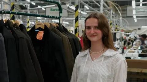 Hannah Karpel/BBC young woman standing in the foreground beside a clothing rail of dark waxed jackets on wooden Barbour hangers. The person is wearing a light striped shirt, hoop earrings and a necklace, and is facing the camera. The setting is a factory or workshop, with industrial lighting, metal framework and workstations visible in the background. Barbour signs can be seen above the clothing rail, and one jacket has an orange tag attached to it. No bags, bins or floor markers are clearly visible.