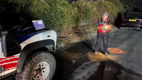 Suffolk Lowland Search and Rescue A woman stands on a road at night time. She is looking at a drone controller with a screen on it. She has short grey hair and is wearing black clothing with a red bib over the top. There are vehicles parked on the front in front and behind her.