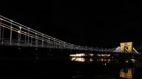 It is night time and a dimly lit suspension bridge crossing a river can be seen. The bridge viaduct can be seen illuminated at one end towards the background.