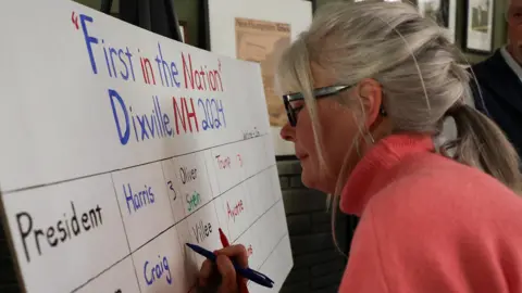 A woman in a pink jumper writes the results of Dixville Notch's vote on a whiteboard.