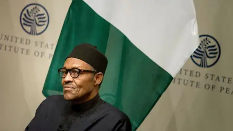 AFP via Getty Images Muhammadu Buhari sits in front of a Nigerian flag and signs that say United States Institute of Peace. He wears a black top, black cap and glasses.