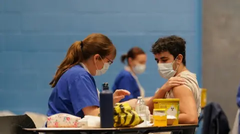 A nurse in scrubs and a mask prepares a vaccination at a table. A patient wearing a face mask has rolled up his sleeve.