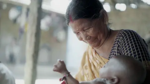 Siro, who has an elderly wizened face, and dressed in a yellow sari, smiles down lovingly at a baby in her arms