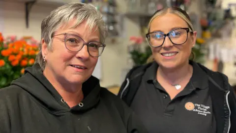 Two women smile. They are florists. Behind them are loads of flowers. 