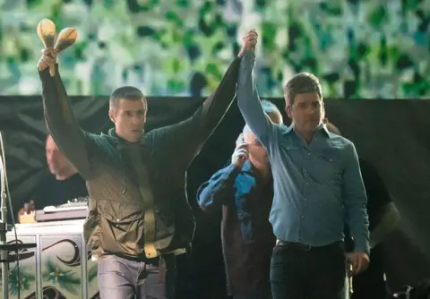 Photograph of Liam and Noel Gallagher standing hand in hand on stage.