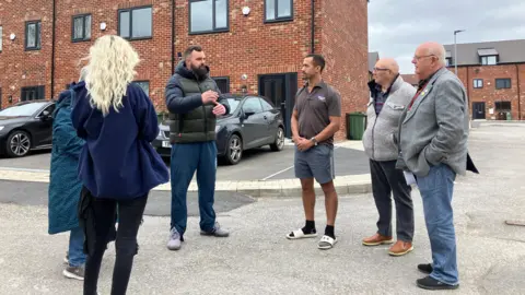 A group of six people stand in a road chatting, new houses are behind them. 