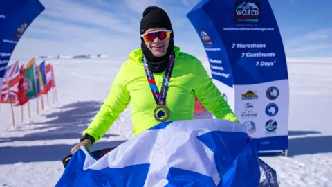World Marathon Challenge Bobby pictured wearing a florescent yellow top, a black woolen snood and sunglasses. He is wearing a medal and holding a saltire flag with a finish line in the background.