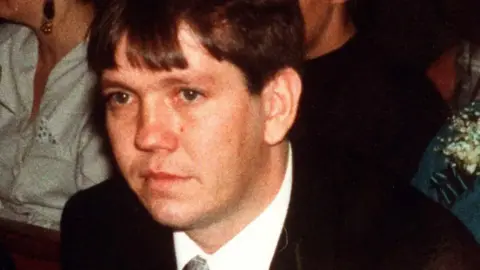 A man wearing a suit and tie looking off camera. There are other people whose faces cannot be seen sitting behind him.