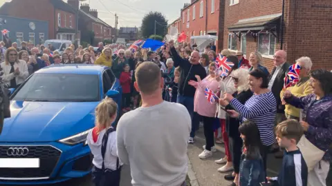 James Burridge/BBC Scores of people in the street waving bunting and Union Jack flags as Maise arrives home in a blue car