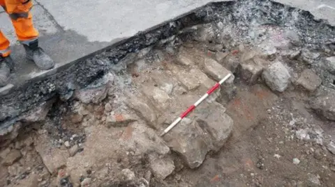 An image of a large hole dug into a concrete road, exposing rubble and bricks below the surface. A red and white striped stick marks out a section of the exposed bricks. To the left, stood on the concrete a pair of legs can be seen, wearing black work boots and florescent orange trousers. 