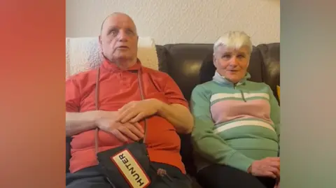 Man with red polo shirt and dark trousers has a bag hanging from his neck and placed on his lap with Hunter written on it. He is sat on a brown sofa with a woman with a patterned green top and dark trousers. There is a beige wall.
