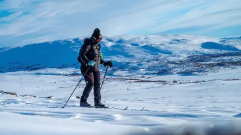 Anya Eames Anya skiing in the arctic. She has full thermal gear and ski poles. A large snowy hill looms behind.