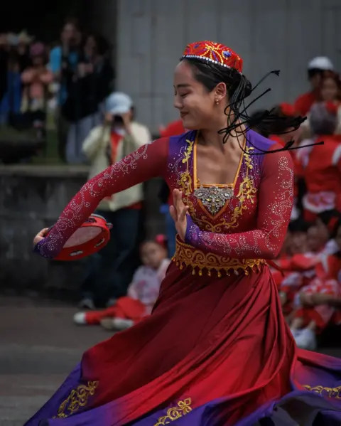 Alyson Macdonald A woman dressed in a red dress with purple and gold on it dances in front of a watching crowd 