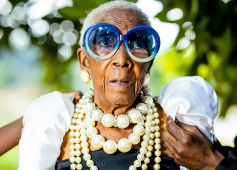 Luxury Media Zambia A hand can be seen adjusting one of Margret Chola's puffy white sleeves. She wears large sunglasses with green frames and four strings of pearls of various sizes