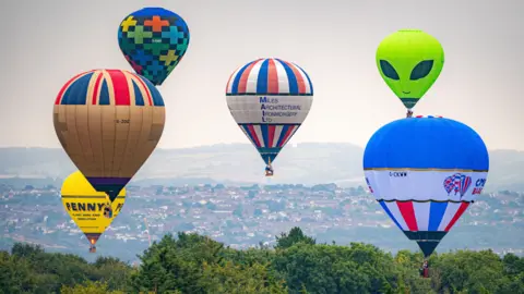 Six hot air balloons in the sky over Bristol. The city can be seen in the distance. The balloons all vary in shape and size. There is a green one shaped like an alien, two blue, red and white striped balloons, one with a union jack print on top, one multicoloured balloon and a yellow one. 