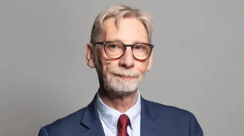 The official portrait of Sir George Howarth. He smiles into the camera, weating black glasses. He has gray, combed-over hair and a short beard. He wears a red tie. 