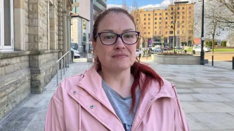 Sinead Quinn with long red hair, in a ponytail, wearing black glasses, a grey t-shirt and a pink trench style jacket. She is stood outside numerous buildings, with higher-rise developments in the background