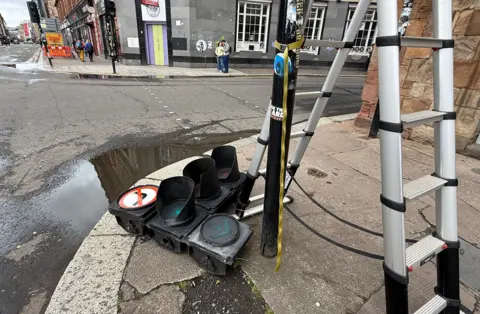 A damaged set of traffic lights lies on the pavement beside a road in Glasgow, with a ladder strapped to a nearby pole and debris visible.