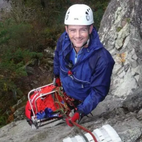 Karl Lester A man smiling a the camera, he is on the mountain side. He has a blue jacket on and a white helmet.