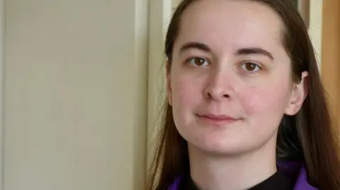 Mollie Cole‑Wilkin looks at the camera. She is wearing a purple uniform with white piping around the edge. She has a black turtle neck top on underneath it. Mollie is standing in the kitchen of Stephen's house. You can see her head and shoulders and there is a wooden kitchen cabinet in the background.