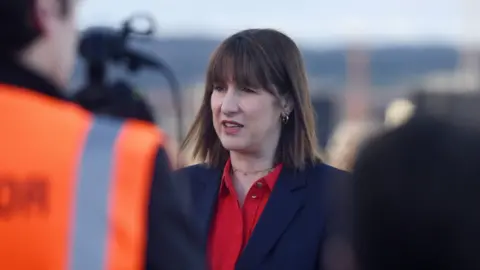 La canciller de los medios de Pensilvania, Rachel Reeves, habla con los periodistas, con una camisa de cuello abierta de escarlata, una chaqueta de traje azul marino y una cadena de gargantillas dorada, con pequeños pendientes de oro de oro. 