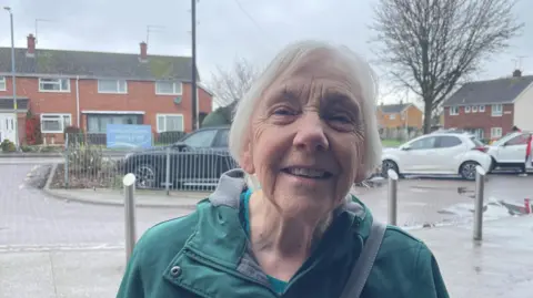 A white haired woman in a green coat is standing on the pavement next to a car park with multiple cars and silver bollards.