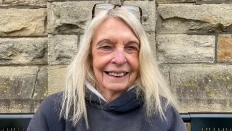 A woman with long, blonde hair and wearing a blue hooded top sitting on a bench with a stone wall behind her.