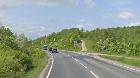 A single lane road with grass verges on either side. A footpath on the right-hand side of the road. Four cars on the road.