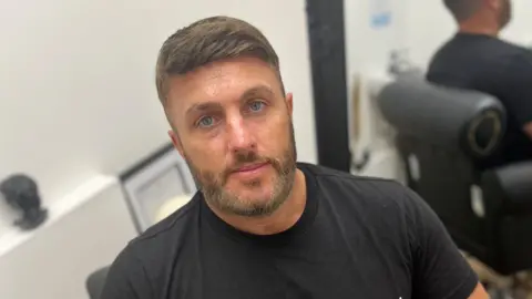 Wayne Brown wearing a wig or hair system, while sitting in a salon. His brown hair is swept to one side.