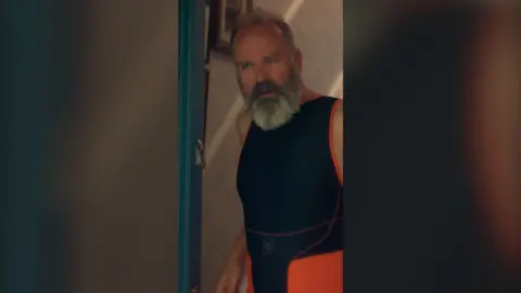 An older man with grey hair and beard wears a sleeveless, black and orange sporting catsuit. He is standing in a front doorway.