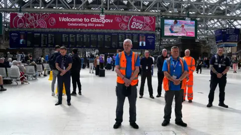 PA Media Men standing wearing orange hi vis vests in front a a train deperture board