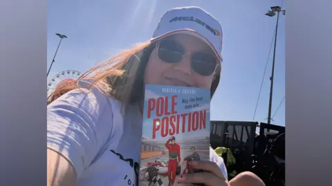 Rebecca Caffery Rebecca is holding her book, Pole Position, in front of her and smiling. She has long auburn hair and is wearing a light blue and orange McLaren cap.
The book has two male formula one drivers on the front, one walking towards a chequered flag and the other sitting on the wheel of a formula one car.