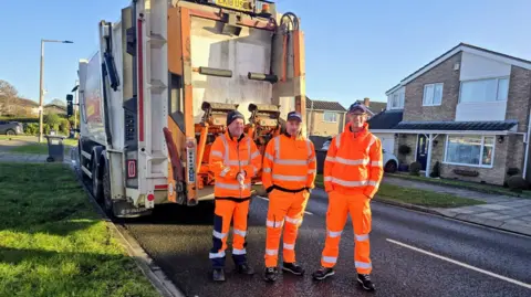 Three refuse workers wearing orange high vis outfits, standing in front of a refuse wagon on a suburban street with the sun shining 