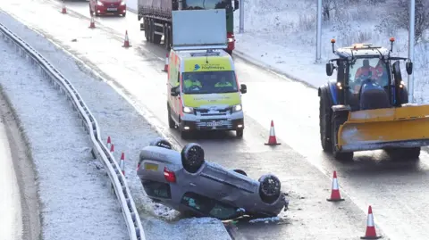 PA Media A grey car overturned on the road which is slick with melted ice. Traffic is being held behind it while a snow plough is parked up alongside it. Snow covers the areas beside the road.