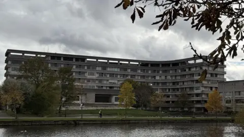 A large, curved concrete building with multiple rows of windows stands behind a grassy area and a pond. The building is Bedford Borough Council's headquarters. Several trees with autumn foliage are scattered across the scene, and branches frame the top of the image. The sky is overcast, creating a moody atmosphere, and a few people can be seen walking along the path near the water.