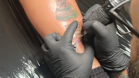 A tattoo artist inks the words Just Joking! under a tattoo of the World Cup and the words England World Cup Winner 2026 on a man's leg