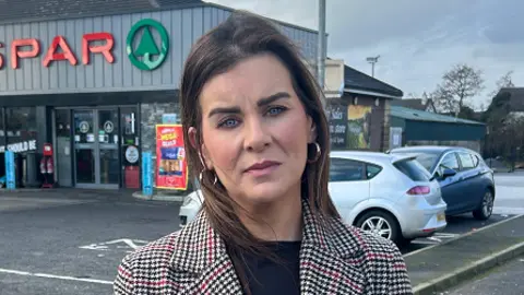 Eimear Carney. She has dark long hair and is wearing a black, red and brown patterned jacket with a black top underneath. She is standing by the SPAR.