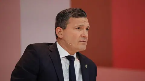 Bloomberg via Getty Images Solomon is photographed at an event, wearing a black suit and tie and a white button-up shirt. He has short black hair and is cleanly shaven. Behind him is a red backdrop of different shades.
