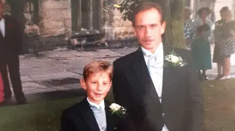 Chris Wild A small boy stands next to his father, a man with dark hair and a side parting. He also has a moustache. Both wear morning suits and it appears they are at a wedding.