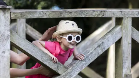 National Trust Images/Annapurna Mellor A young girl with blond hair, a sun hat with a smiley face on the front and white flower style sunglasses pokes her head through a gap on a wooden bridge. She is wearing a hot pink t-shirt and an adult is holding her from behind.
