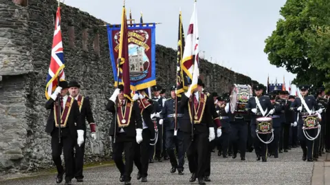 A group of men wearing black suits and black bowler hats marching in a parade. They are wearing maroon sashes an carrying various flags. There is a band behind them and old walls to the left.