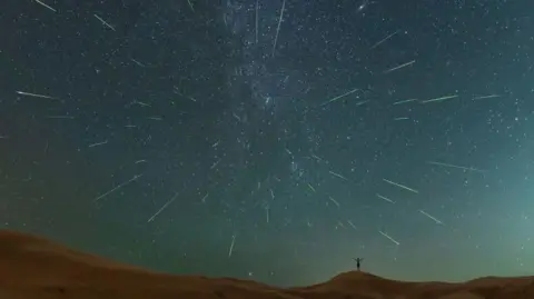 Getty Images A figure stands on the hills at night, a long exposure photo captures several meteors in the night sky.