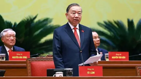 To Lam is delivering a speech at a party session. He is wearing a suit and a red tie.