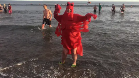 Alan Hughes A cold looking dipper with their arms outstretched in the water wearing a long red lobster costume. 