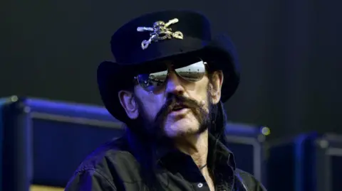 BBC/Steve Barney A head and shoulders shot of the singer in a black shirt, dark glasses and black hat with a silver skull and cross bones on a stage. He has a dark handlebar moustache.