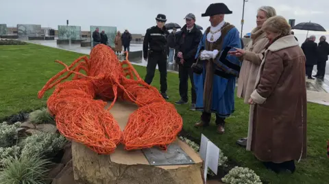 The sculpture is srrounded by a group of people who are examining it. It sits on a stone plinth surrounded by planting and the wire lobst is bright orange.