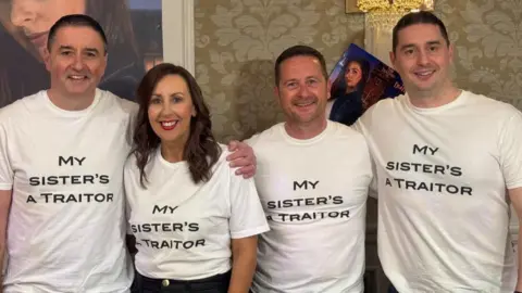 Three men and one woman are arm in arm, looking into the camera and smiling. They are all wearing white tshirts that say 'My sister's a Traitor'.