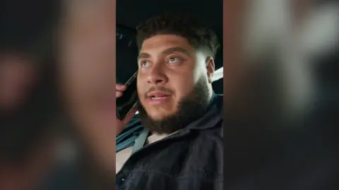 A man, Big Zuu, with short black hair and a beard, talks into a phone while sat in a car