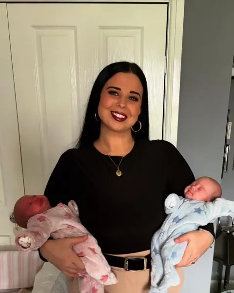 Katie Matthews The picture shows a woman smiling at the camera. She's wearing a black long sleeved top and beige trousers. She has black hair and is wearing hooped earrings. She's holding a new born baby in each arm. One is wearing a pink baby grow, and the other is wearing a blue baby grow.